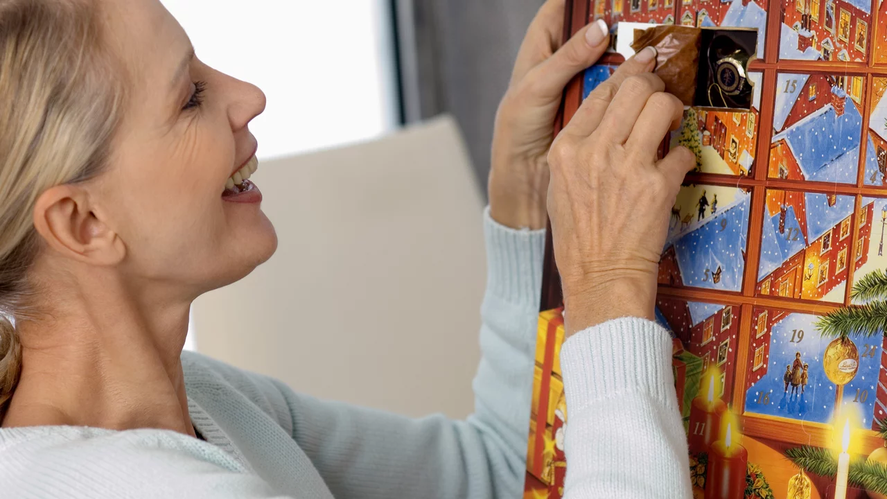 Eine lächelnde Frau öffnet ein Türchen eines Adventskalenders. Der Kalender zeigt weihnachtliche Motive. Die Szene strahlt Freude und Vorfreude auf das Weihnachtsfest aus.