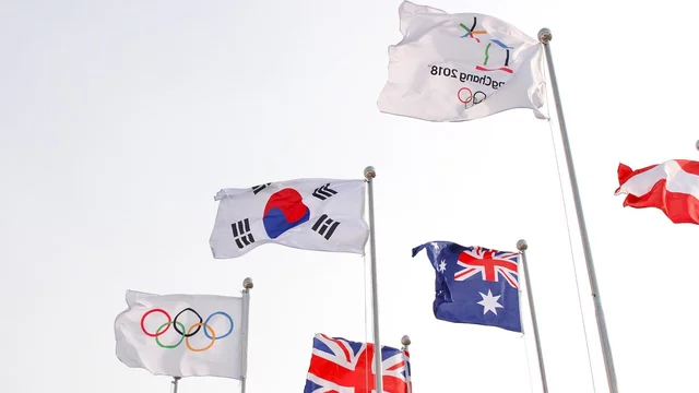Eine Reihe von Fahnen weht in der Luft. Darunter befinden sich nationale Fahnen, einschließlich der Südkoreanischen, sowie die Olympischen Ringe. Die Fahnen symbolisieren die Teilnahme verschiedener Länder an den Olympischen Spielen.