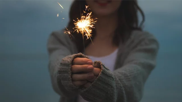Eine Frau hält eine brennende Wunderkerze in der Hand. Die Funken sprühen hervor und erzeugen ein festliches Licht. Der Hintergrund zeigt einen dämmerigen Himmel über Wasser, was eine stimmungsvolle Atmosphäre schafft.