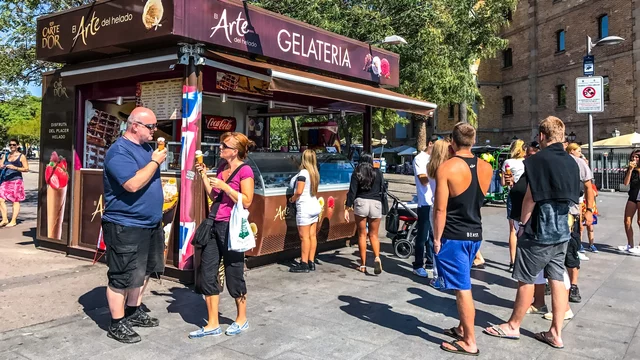 Ein Eisstand mit der Aufschrift "Gelateria" ist umgeben von einer Warteschlange von Personen. Einige Gäste halten Eis in der Hand, während andere auf ihre Bestellung warten. Im Hintergrund sind Bäume und Gebäude sichtbar.