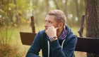 Ein nachdenklicher Mann sitzt im Freien auf einer Bank, umgeben von Bäumen und sanften Lichtverhältnissen. Er hat einen blauen Kapuzenpullover und schaut in die Ferne, während er mit der Hand sein Gesicht stützt.