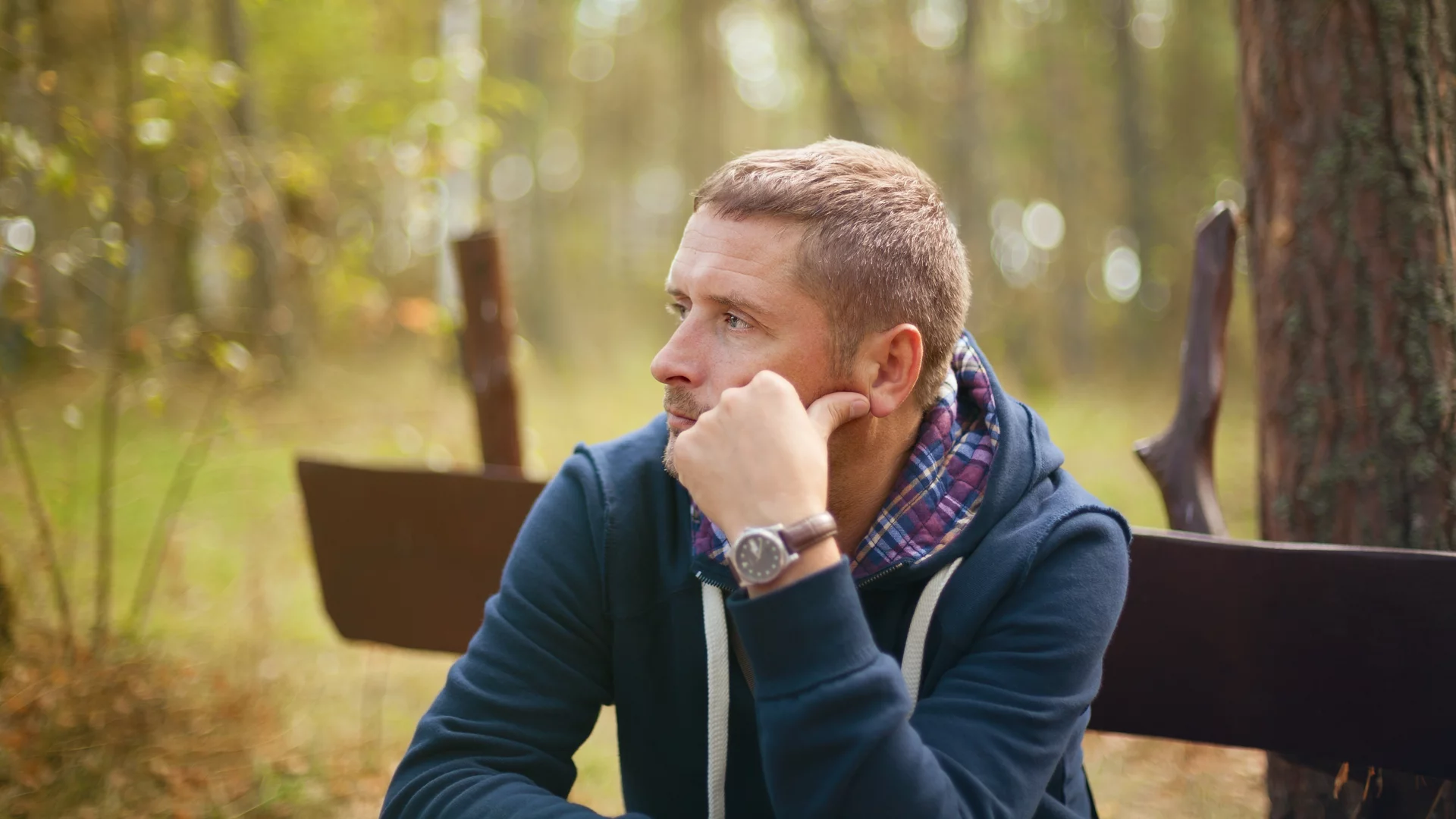 Ein nachdenklicher Mann sitzt im Freien auf einer Bank, umgeben von Bäumen und sanften Lichtverhältnissen. Er hat einen blauen Kapuzenpullover und schaut in die Ferne, während er mit der Hand sein Gesicht stützt.