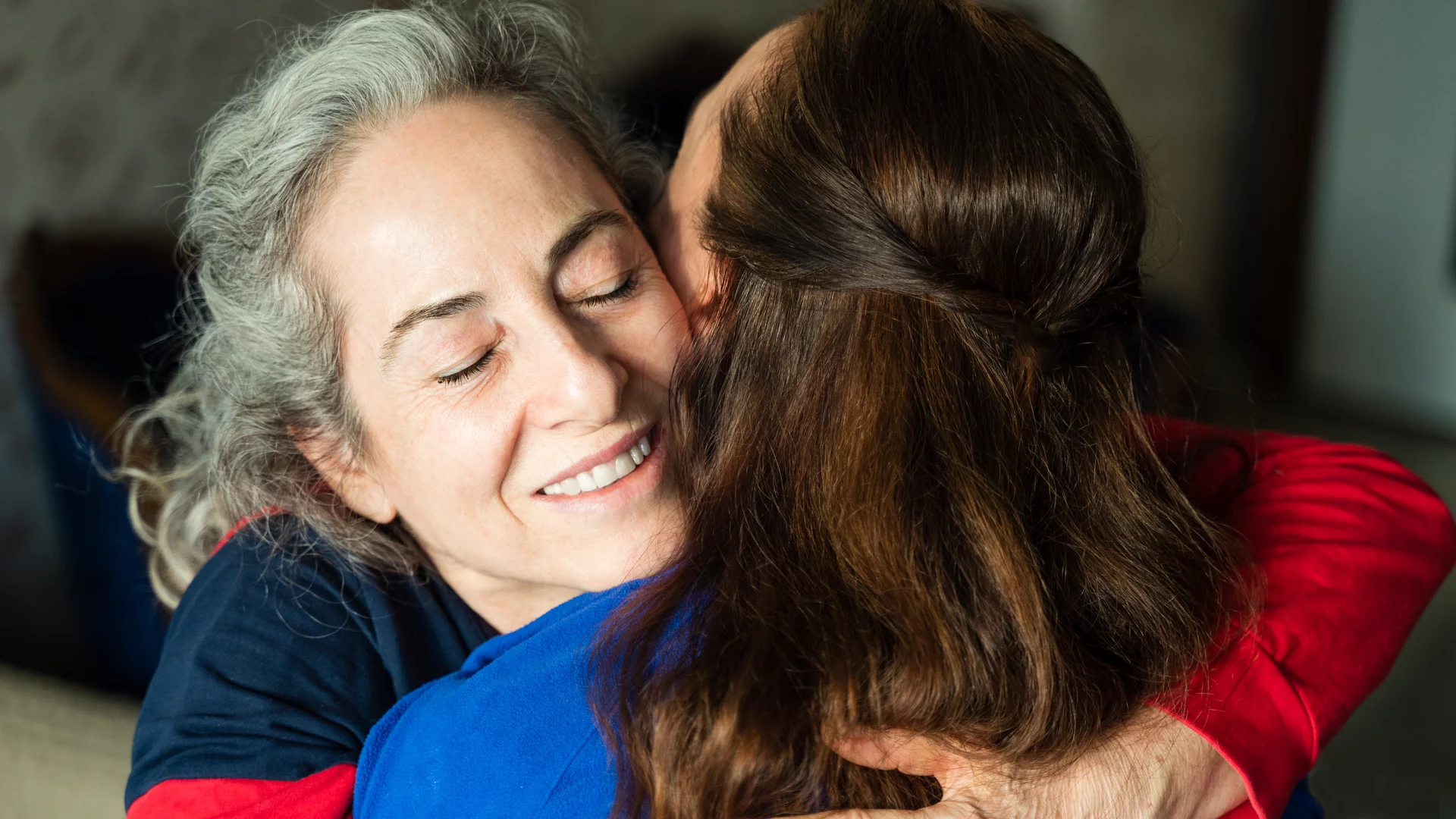 Zwei Frauen umarmen sich herzlich. Die ältere Frau mit grauen Haaren lächelt sanft, während sie ihre Augen schließt. Die jüngere Frau, mit langen braunen Haaren, drückt sie liebevoll. Die Szene vermittelt Wärme und Zuneigung zwischen den beiden.