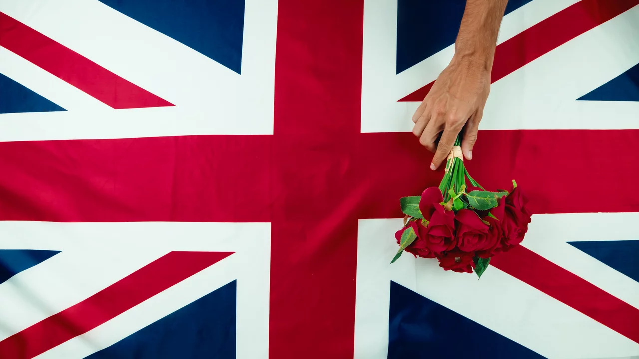 Eine Hand hält einen Strauß roter Rosen, der auf einer Flagge des Vereinigten Königreichs liegt. Die Flagge zeigt den typischen roten, weißen und blauen Hintergrund. Die Szene vermittelt einen romantischen und nationalen Bezug.