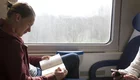 Ein junger Mann sitzt in einem Zug und liest ein Buch. Er trägt einen roten Hoodie und schaut konzentriert auf die Seite. Neben ihm sitzt eine Person, die ein Smartphone in der Hand hält. Im Hintergrund ist ein Fenster mit verschwommenem Blick auf die Landschaft.