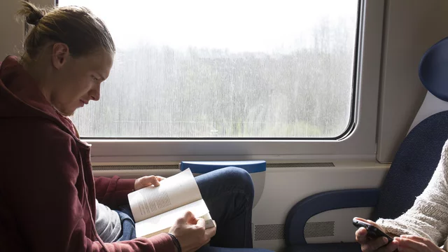 Ein junger Mann sitzt in einem Zug und liest ein Buch. Er trägt einen roten Hoodie und schaut konzentriert auf die Seite. Neben ihm sitzt eine Person, die ein Smartphone in der Hand hält. Im Hintergrund ist ein Fenster mit verschwommenem Blick auf die Landschaft.