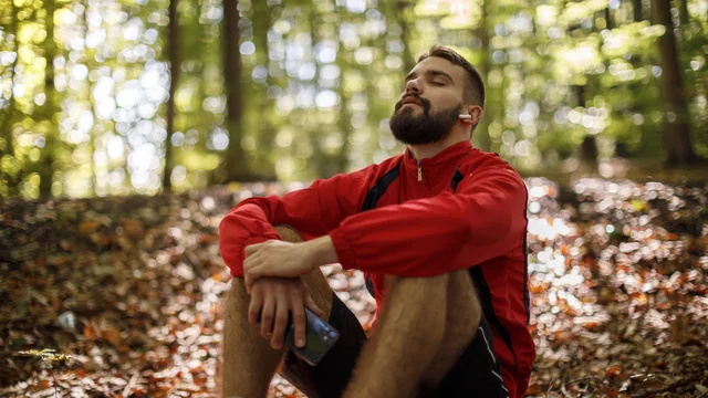 Ein Mann sitzt auf dem Boden im Wald, umgeben von Laub. Er trägt ein rotes Sportjackett und Kopfhörer. Mit geschlossenen Augen genießt er die Ruhe und scheint entspannt. In seiner Hand hält er eine Getränkeflasche. Sonnenlicht strahlt durch die Bäume.
