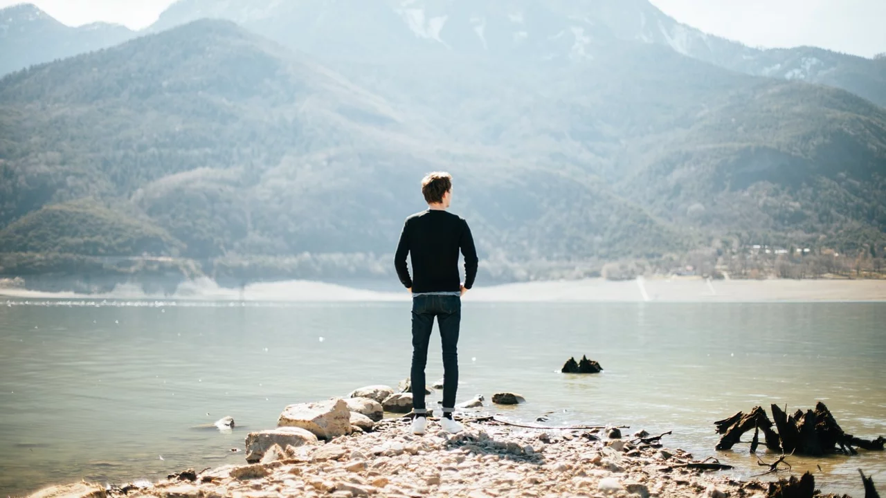 Eine Person steht auf einem Steg am Ufer eines ruhigen Gewässers und blickt auf eine beeindruckende Berglandschaft im Hintergrund. Die Szenerie vermittelt ein Gefühl von Ruhe und Weite in der Natur.