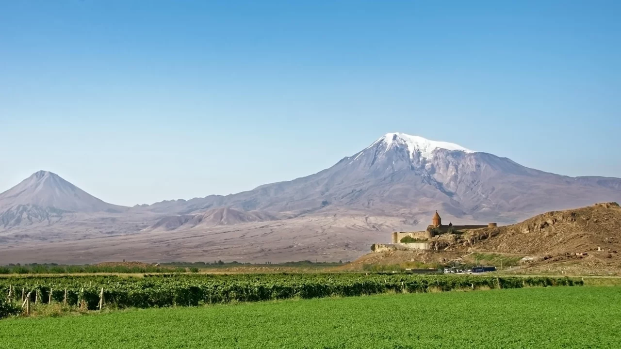 Die Aufnahme zeigt die majestätischen Vulkane Ararat und seine kleinere Schwester in einer weitläufigen Landschaft. Im Vordergrund erstreckt sich ein grünes Feld, während die schneebedeckten Gipfel der Berge im Hintergrund deutlich sichtbar sind.