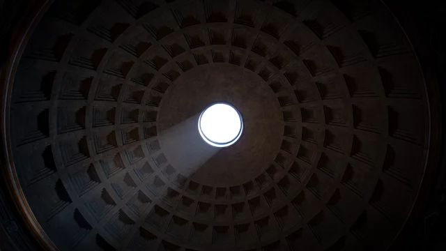 Das Bild zeigt die Decke eines großen, runden Raums mit einer auffälligen Öffnung in der Mitte. Lichtstrahlen fallen durch das Loch und erzeugen eine helle, lebendige Szene, während die geometrischen Muster der Decke den Raum umrahmen.