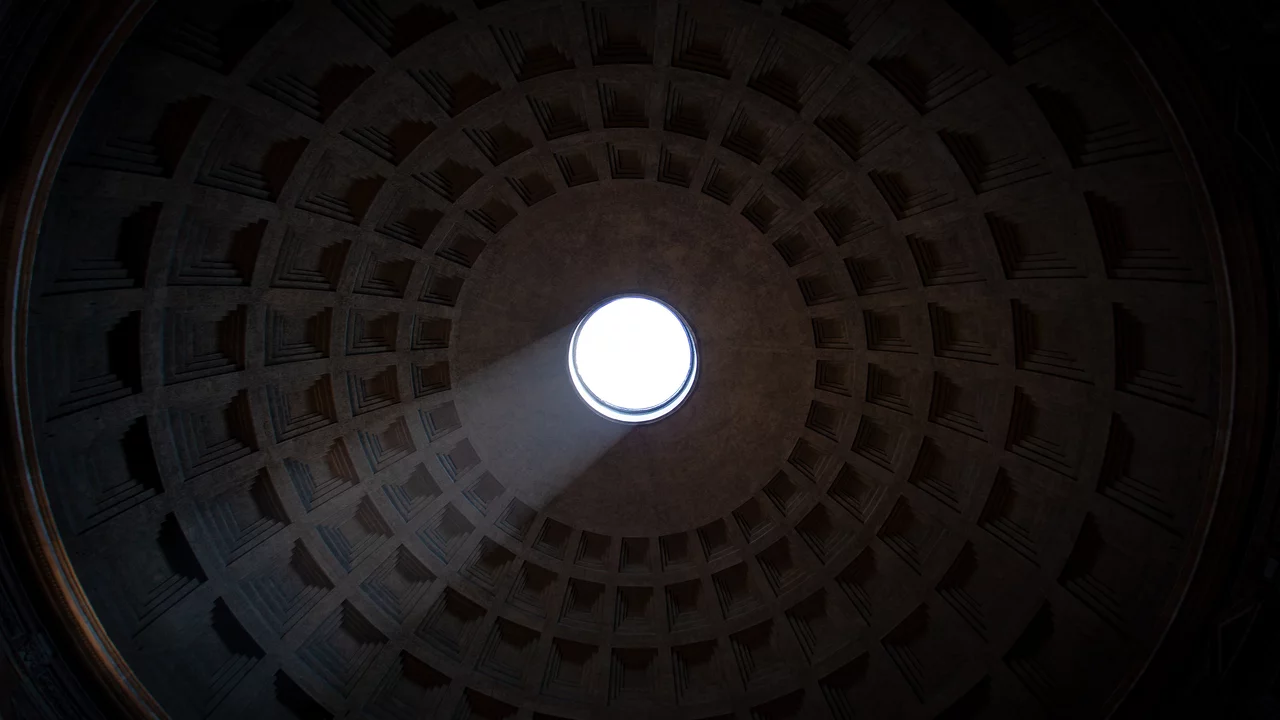 Das Bild zeigt die Decke eines großen, runden Raums mit einer auffälligen Öffnung in der Mitte. Lichtstrahlen fallen durch das Loch und erzeugen eine helle, lebendige Szene, während die geometrischen Muster der Decke den Raum umrahmen.