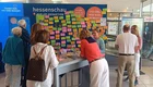 Gruppengespräch vor einer Wand voller bunter Haftnotizen in einem Pop-up-Bereich der Hessenschau. Die Menschen diskutieren, schreiben und teilen ihre Gedanken. Das Motto „Sag uns, was dich bewegt“ ist deutlich sichtbar.