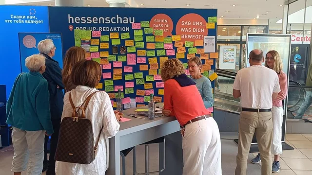 Gruppengespräch vor einer Wand voller bunter Haftnotizen in einem Pop-up-Bereich der Hessenschau. Die Menschen diskutieren, schreiben und teilen ihre Gedanken. Das Motto „Sag uns, was dich bewegt“ ist deutlich sichtbar.