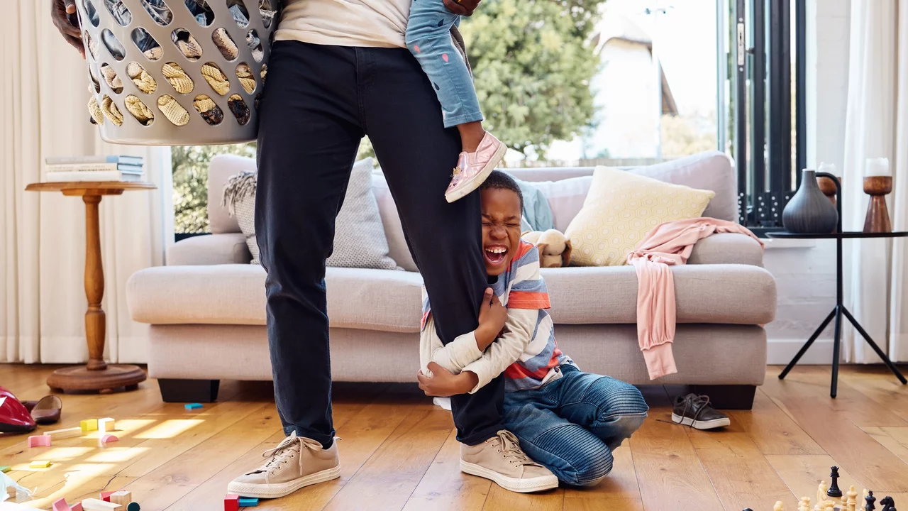 Ein Erwachsener trägt einen Wäschekorb und hält ein Kind an seinem Bein, während ein weiteres Kind lachend auf dem Boden sitzt. Die Szene ist in einem unordentlichen Wohnzimmer mit Spielzeug und Möbeln im Hintergrund dargestellt.