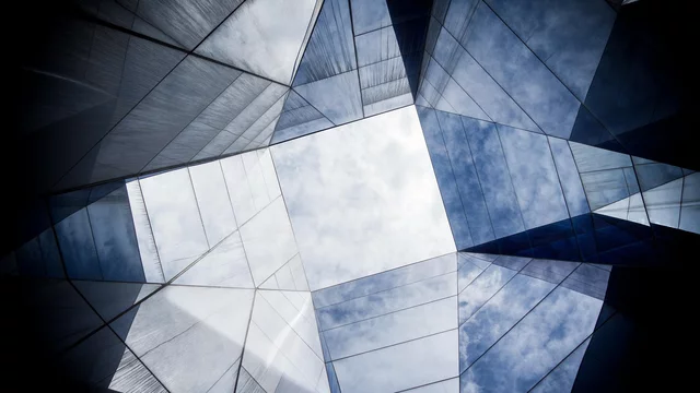 Blick auf den Himmel durch eine geometrische Struktur aus spiegelnden Flächen. Die verschiedenen Materialien reflektieren Licht und erzeugen ein faszinierendes Muster. Der Himmel zeigt sich in einem klaren, hellen Bereich zwischen den schrägen, lichtdurchlässigen Elementen.