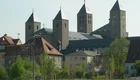 Eine historische Kirche mit mehreren Türmen und spitzen Dächern, umgeben von Wohnhäusern und Bäumen. Die Architektur ist aus hellem Stein, und die Türme sind mit kleinen Kreuzen geschmückt. Die Szenerie vermittelt einen friedlichen Eindruck.