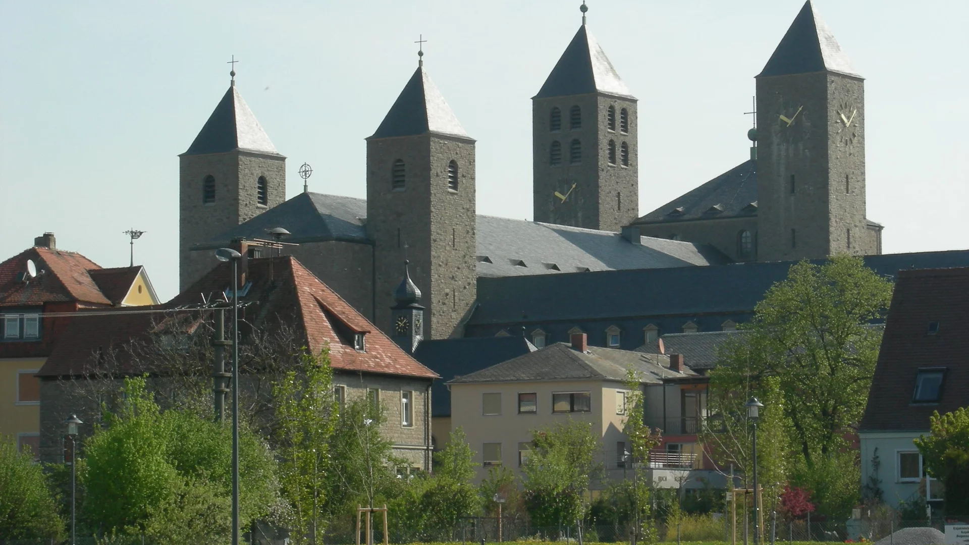 Eine historische Kirche mit mehreren Türmen und spitzen Dächern, umgeben von Wohnhäusern und Bäumen. Die Architektur ist aus hellem Stein, und die Türme sind mit kleinen Kreuzen geschmückt. Die Szenerie vermittelt einen friedlichen Eindruck.