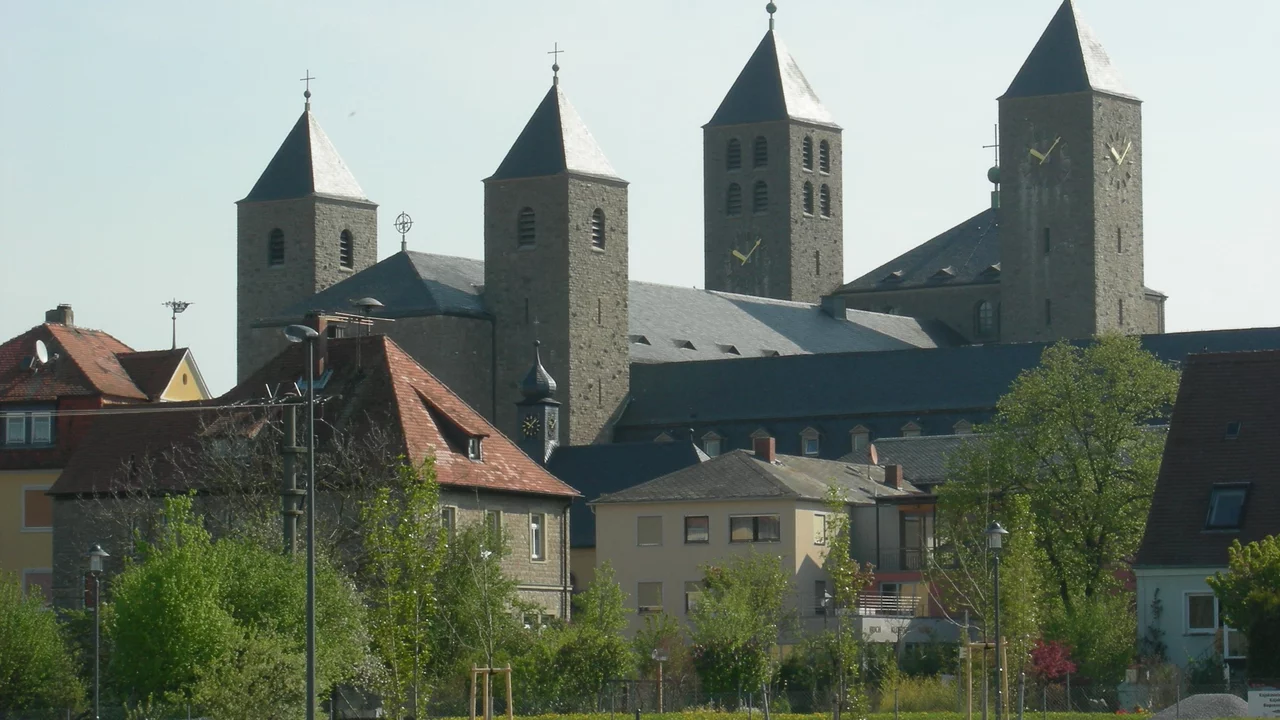 Eine historische Kirche mit mehreren Türmen und spitzen Dächern, umgeben von Wohnhäusern und Bäumen. Die Architektur ist aus hellem Stein, und die Türme sind mit kleinen Kreuzen geschmückt. Die Szenerie vermittelt einen friedlichen Eindruck.