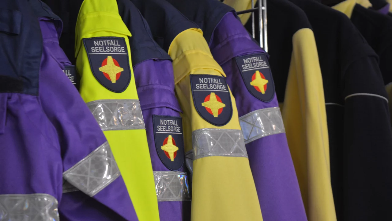 Mehrere Einsatzjacken sind auf einem Kleiderständer hängen. Die Jacken sind in lila und gelb gehalten und tragen das Emblem der Notfallseelsorge. Die reflektierenden Streifen an den Jacken sind gut sichtbar, was auf ihre Funktion im Einsatz hinweist.