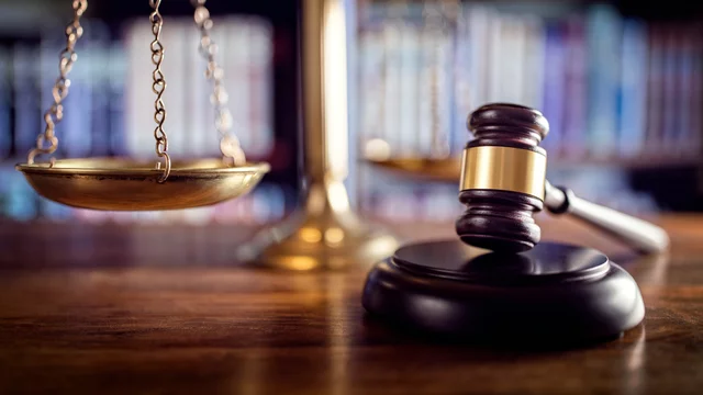 Das Bild zeigt eine Waage der Gerechtigkeit und einen Richterhammer auf einem Holztisch. Im Hintergrund sind unscharfe Bücherregale zu erkennen. Die Waage symbolisiert Gerechtigkeit, während der Hammer für die Autorität des Rechts steht.