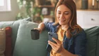 Eine lächelnde Frau sitzt auf einem Sofa und hält eine Tasse in der Hand. Sie blickt auf ihr Smartphone und wirkt entspannt. Im Hintergrund sind Pflanzen und Bücherregale sichtbar, die eine gemütliche Atmosphäre schaffen.