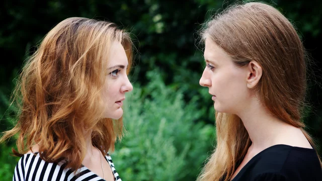 Zwei Frauen stehen sich gegenüber und schauen sich intensiv in die Augen. Eine Frau hat lockiges, blondes Haar und trägt ein gestreiftes Oberteil, während die andere Frau langes, glattes, braunes Haar hat und ein schwarzes Oberteil trägt. Der Hintergrund ist grün und verschwommen.