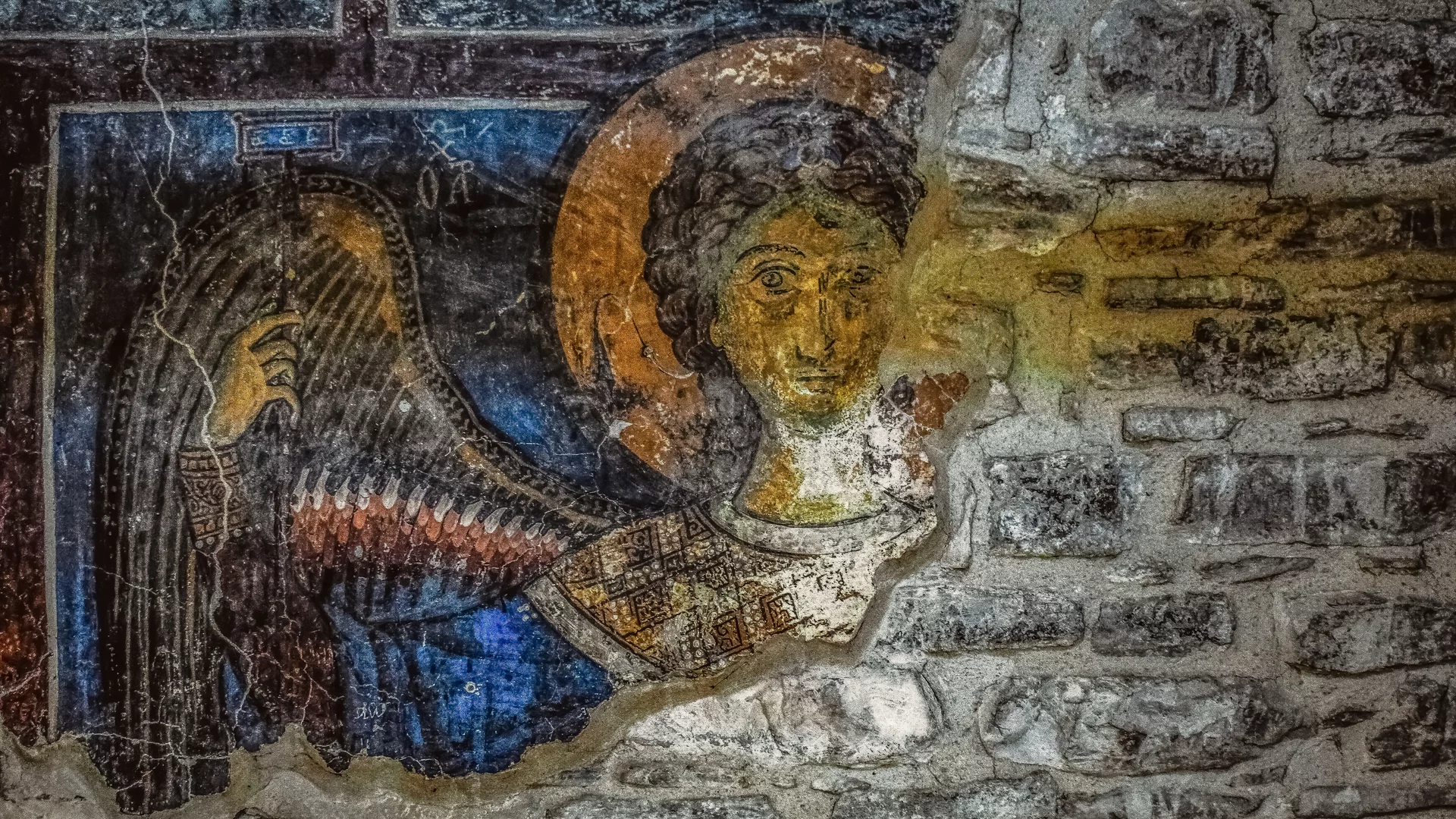 Das Bild zeigt ein Wandgemälde eines Engels mit Flügeln und einem Heiligenschein. Der Engel ist frontal abgebildet und scheint nach vorne zu schauen. Die Farben sind überwiegend Blau, Gold und Beige, wobei die Mauer im Hintergrund aus unregelmäßigen Steinen besteht.