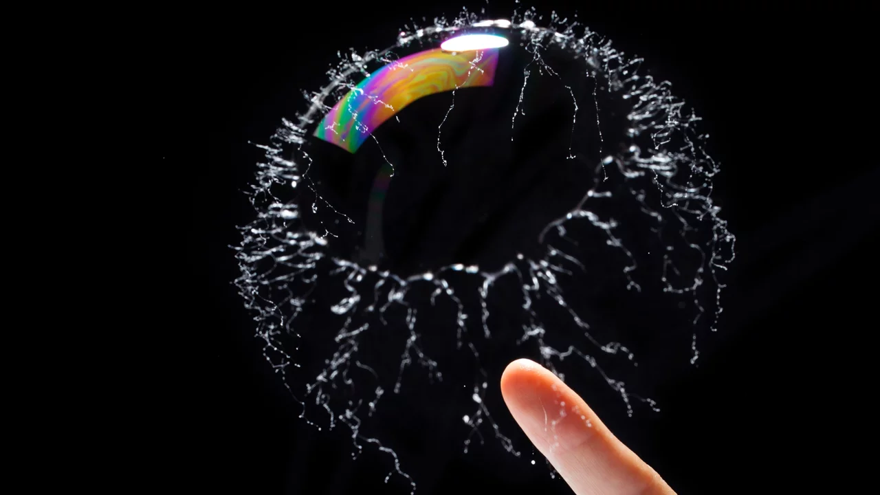 Eine Seifenblase schwebt vor einem schwarzen Hintergrund. Ein Finger nähert sich der Blase, die von feinen Wassertröpfchen umgeben ist. Die Blase zeigt einen schimmernden Regenbogen-Effekt.