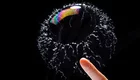 Eine Seifenblase schwebt vor einem schwarzen Hintergrund. Ein Finger nähert sich der Blase, die von feinen Wassertröpfchen umgeben ist. Die Blase zeigt einen schimmernden Regenbogen-Effekt.