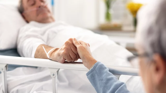 Eine Person im Krankenhausbett hält die Hand einer anderen Person. Der Patient wirkt schwach, erhält jedoch Unterstützung und Trost durch den Händedruck. Die Szene vermittelt Gefühle von Nähe und Fürsorge.