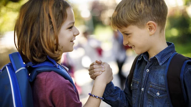 Zwei Kinder, ein Mädchen und ein Junge, stehen sich gegenüber und halten sich an den Händen. Sie lächeln einander an, während sie die Hände schütteln. Im Hintergrund sind unscharfe Figuren zu sehen, die auf einem Weg stehen. Die Szene vermittelt Freude und Freundschaft.