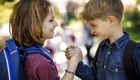 Zwei Kinder, ein Mädchen und ein Junge, stehen sich gegenüber und halten sich an den Händen. Sie lächeln einander an, während sie die Hände schütteln. Im Hintergrund sind unscharfe Figuren zu sehen, die auf einem Weg stehen. Die Szene vermittelt Freude und Freundschaft.
