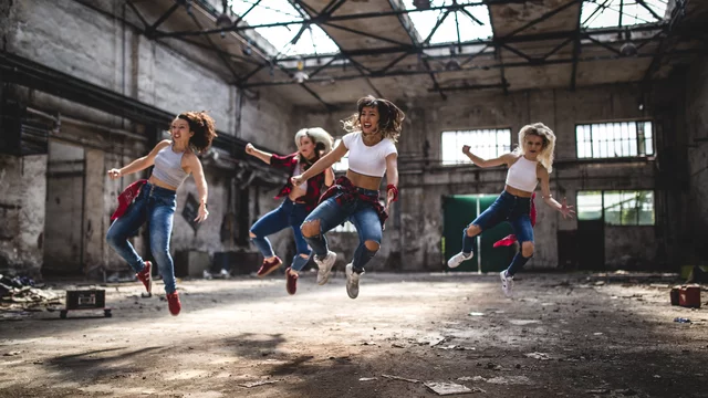 Fünf Frauen in sportlicher Kleidung springen gleichzeitig in einem alten, ungenutzten Raum mit hohen Decken. Der Boden ist uneben und es sind einige Kisten sichtbar. Die Atmosphäre ist energetisch und dynamisch.