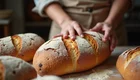 Eine Person mit einem Holzbrett drückt sanft auf frisch gebackenes Brot. Um sie herum liegen mehrere Laibe Brot. 