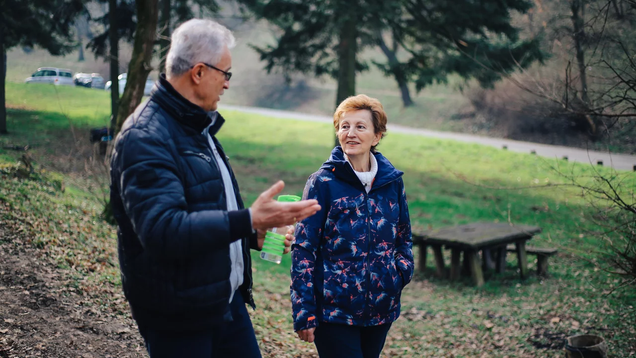 Zwei Menschen, ein älterer Mann und eine ältere Frau, unterhalten sich lächelnd in einem parkähnlichen Gelände mit Bäumen im Hintergrund. Der Mann gestikuliert mit seiner Hand, während die Frau aufmerksam zuhört.
