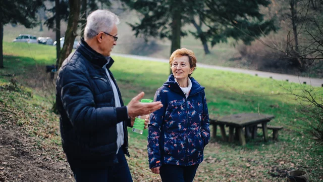 Zwei Menschen, ein älterer Mann und eine ältere Frau, unterhalten sich lächelnd in einem parkähnlichen Gelände mit Bäumen im Hintergrund. Der Mann gestikuliert mit seiner Hand, während die Frau aufmerksam zuhört.