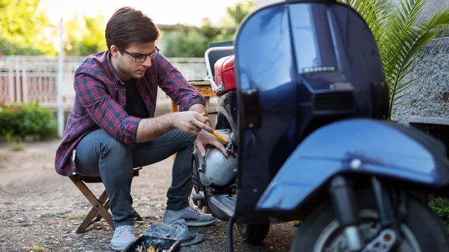 Ein junger Mann in einem karierten Hemd sitzt neben einem Roller und arbeitet daran. Er verwendet Werkzeug, um den Motor zu reparieren. Der Hintergrund zeigt eine sonnige Umgebung mit Pflanzen.