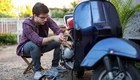 Ein junger Mann in einem karierten Hemd sitzt neben einem Roller und arbeitet daran. Er verwendet Werkzeug, um den Motor zu reparieren. Der Hintergrund zeigt eine sonnige Umgebung mit Pflanzen.
