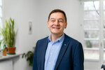 Ein lächelnder Mann in formeller Kleidung steht in einem hellen Raum mit großen Fenstern. Im Hintergrund sind Pflanzen sichtbar. Der Mann trägt ein blaues Jackett über einem karierten Hemd und hat ein Kreuzanhänger an der Halskette.