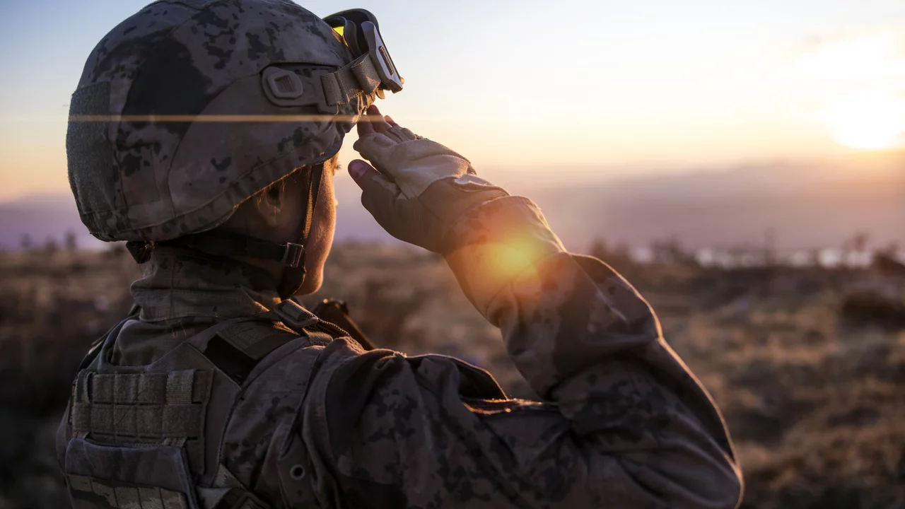 Ein Soldat in Tarnuniform steht im Sonnenuntergang und salutiert. Der Hintergrund zeigt eine weite, unebene Landschaft. Die Sonne strahlt warmes Licht, das den Soldaten und die Szenerie beleuchtet.