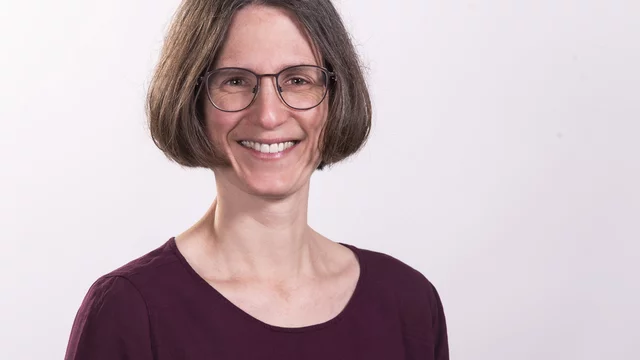 Porträt einer Frau mit schulterlangen, glatten Haaren und einer Brille. Sie trägt ein lila Oberteil und lächelt freundlich in die Kamera. Der Hintergrund ist hell und neutral.