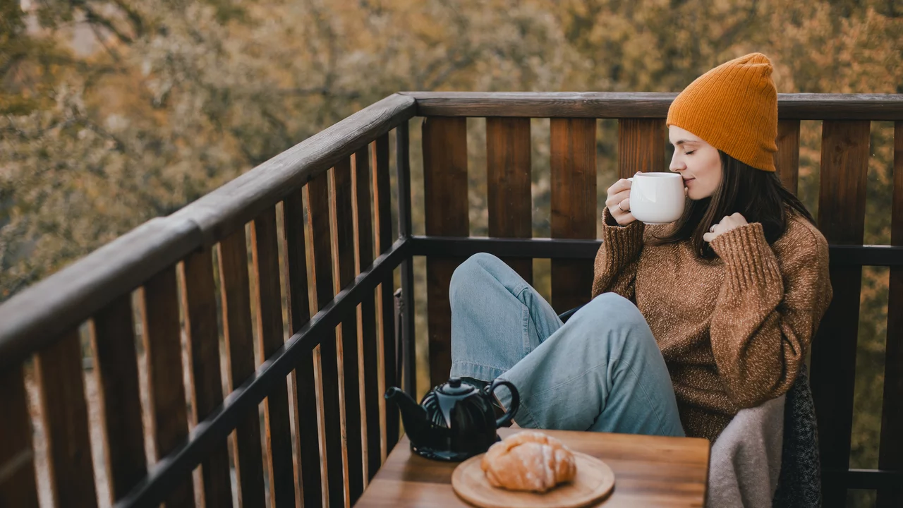 Eine junge Frau mit einer orangefarbenen Mütze sitzt auf einem Balkon, hält eine Tasse in der Hand und genießt eine Aussicht auf herbstliche Bäume. Vor ihr auf einem kleinen Tisch liegt ein Croissant neben einer Teekanne. Die Frau trägt einen gemütlichen Pullover und wirkt entspannt.