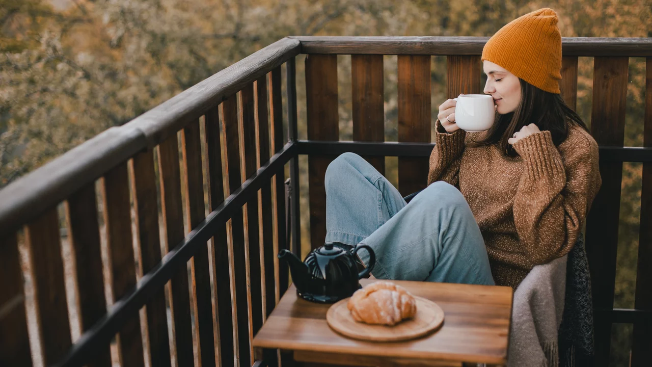 Eine junge Frau mit einer orangefarbenen Mütze sitzt auf einem Balkon, hält eine Tasse in der Hand und genießt eine Aussicht auf herbstliche Bäume. Vor ihr auf einem kleinen Tisch liegt ein Croissant neben einer Teekanne. Die Frau trägt einen gemütlichen Pullover und wirkt entspannt.