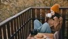 Eine junge Frau mit einer orangefarbenen Mütze sitzt auf einem Balkon, hält eine Tasse in der Hand und genießt eine Aussicht auf herbstliche Bäume. Vor ihr auf einem kleinen Tisch liegt ein Croissant neben einer Teekanne. Die Frau trägt einen gemütlichen Pullover und wirkt entspannt.