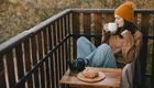 Eine junge Frau mit einer orangefarbenen Mütze sitzt auf einem Balkon, hält eine Tasse in der Hand und genießt eine Aussicht auf herbstliche Bäume. Vor ihr auf einem kleinen Tisch liegt ein Croissant neben einer Teekanne. Die Frau trägt einen gemütlichen Pullover und wirkt entspannt.