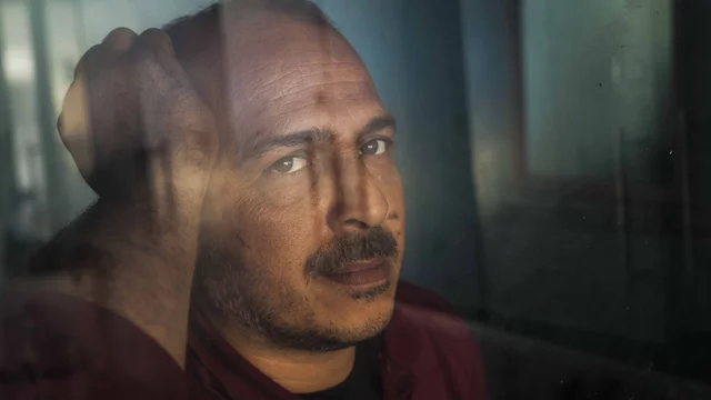 Ein Mann mit kurzen, dunklen Haaren und einem leichten Bart schaut ernst in die Kamera. Er hat eine nachdenkliche Miene und berührt mit einer Hand seinen Kopf. Der Hintergrund wirkt verschwommen, was den Fokus auf sein Gesicht lenkt.