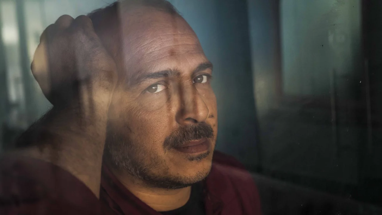 Ein Mann mit kurzen, dunklen Haaren und einem leichten Bart schaut ernst in die Kamera. Er hat eine nachdenkliche Miene und berührt mit einer Hand seinen Kopf. Der Hintergrund wirkt verschwommen, was den Fokus auf sein Gesicht lenkt.