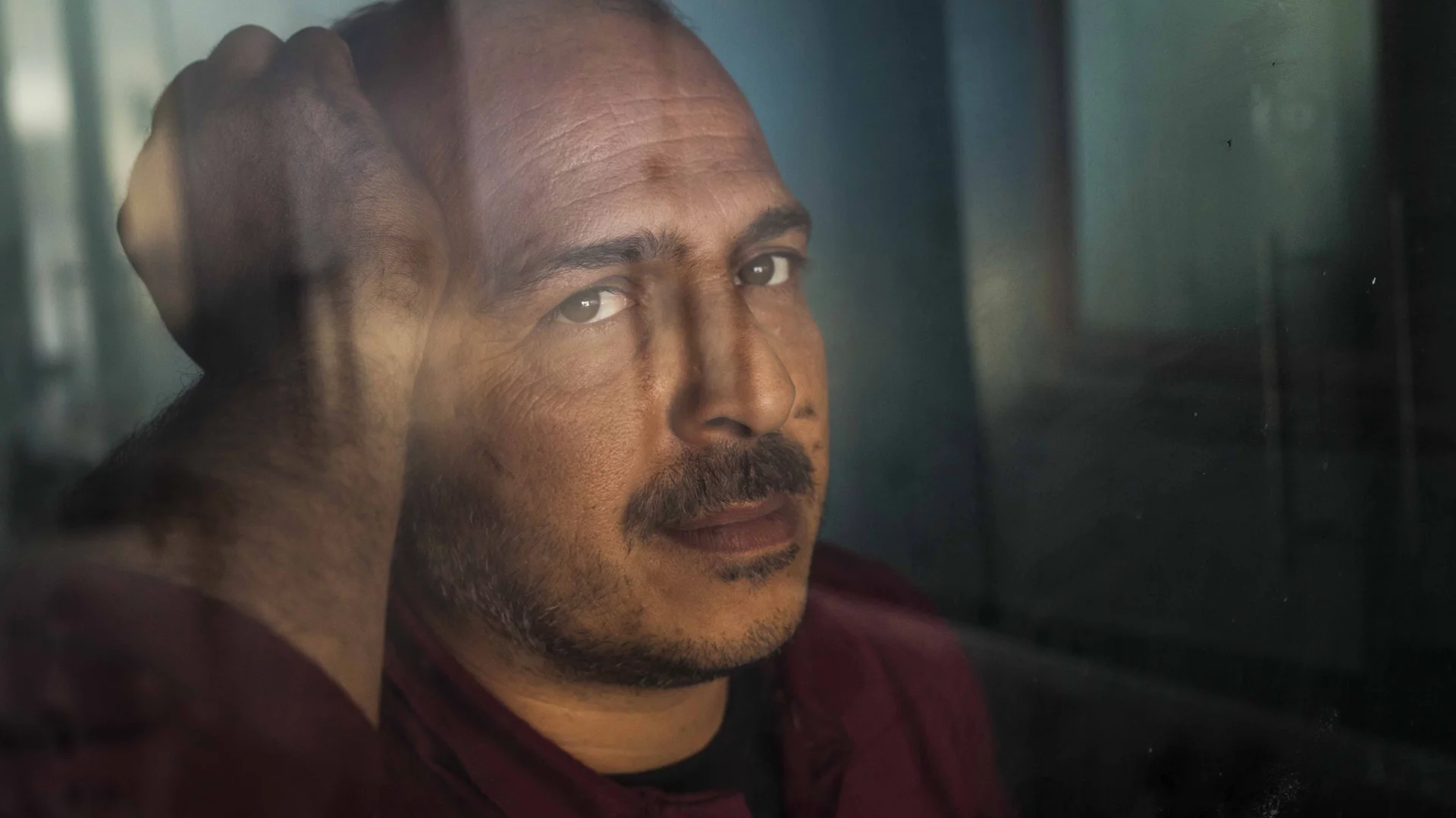 Ein Mann mit kurzen, dunklen Haaren und einem leichten Bart schaut ernst in die Kamera. Er hat eine nachdenkliche Miene und berührt mit einer Hand seinen Kopf. Der Hintergrund wirkt verschwommen, was den Fokus auf sein Gesicht lenkt.