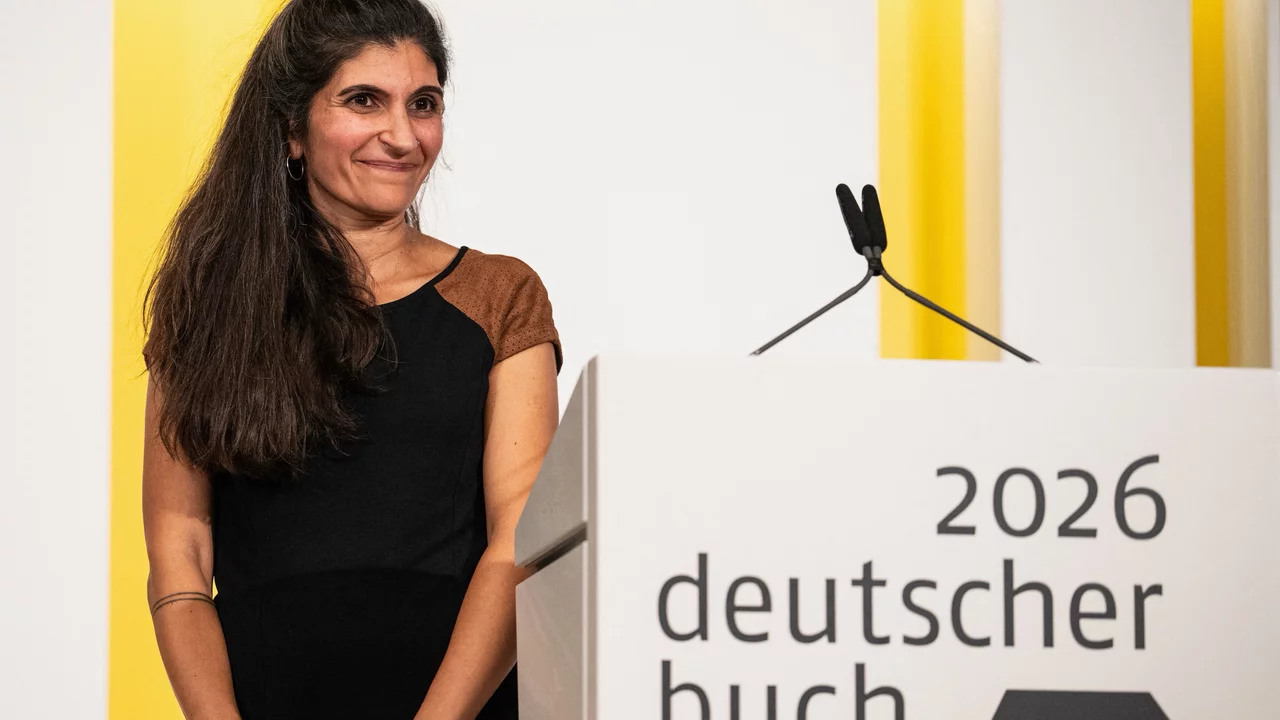 Eine lächelnde Frau mit langen, lockigen Haaren steht neben einem Podest mit der Aufschrift "2026 deutscher buchpreis". Sie trägt ein schwarzes Kleid mit braunen Ärmeln und scheint auf eine Auszeichnung oder eine Ansprache vorbereitet zu sein.