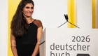 Eine lächelnde Frau mit langen, lockigen Haaren steht neben einem Podest mit der Aufschrift "2026 deutscher buchpreis". Sie trägt ein schwarzes Kleid mit braunen Ärmeln und scheint auf eine Auszeichnung oder eine Ansprache vorbereitet zu sein.